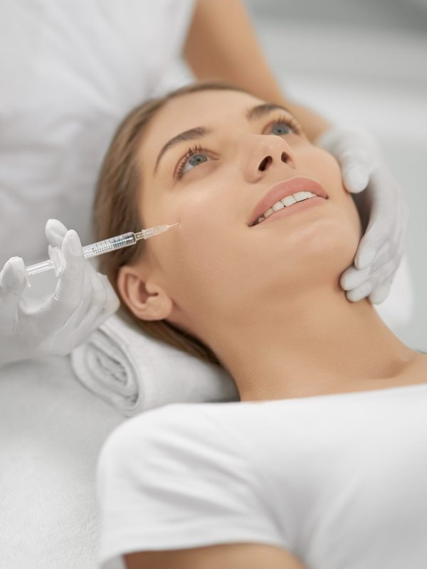Close up of smiling young brunette woman on procedure injection anti-aging skin in beautician. Concept of process injection for improve the condition face.