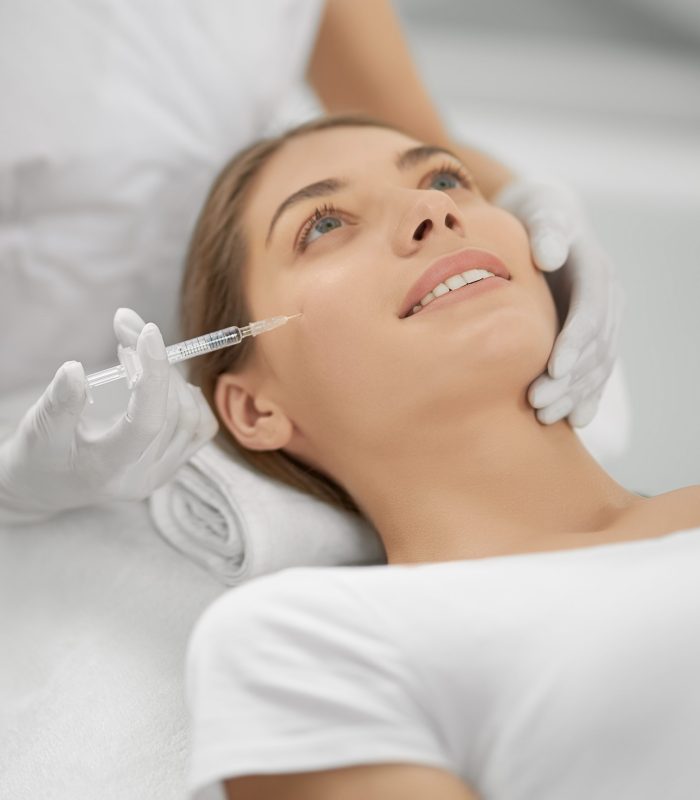 Close up of smiling young brunette woman on procedure injection anti-aging skin in beautician. Concept of process injection for improve the condition face.