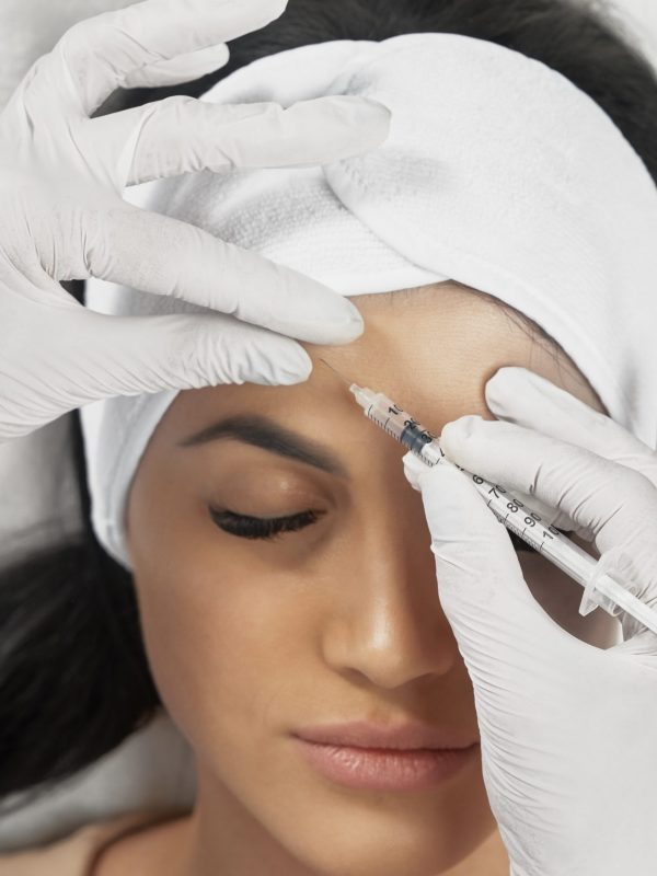 From above view of hyaluronic injection in female forehead. Cosmetologist using syringe with filler while brunette patient with closed eyes, in towel on head lying. Concept of cosmetology, beauty.